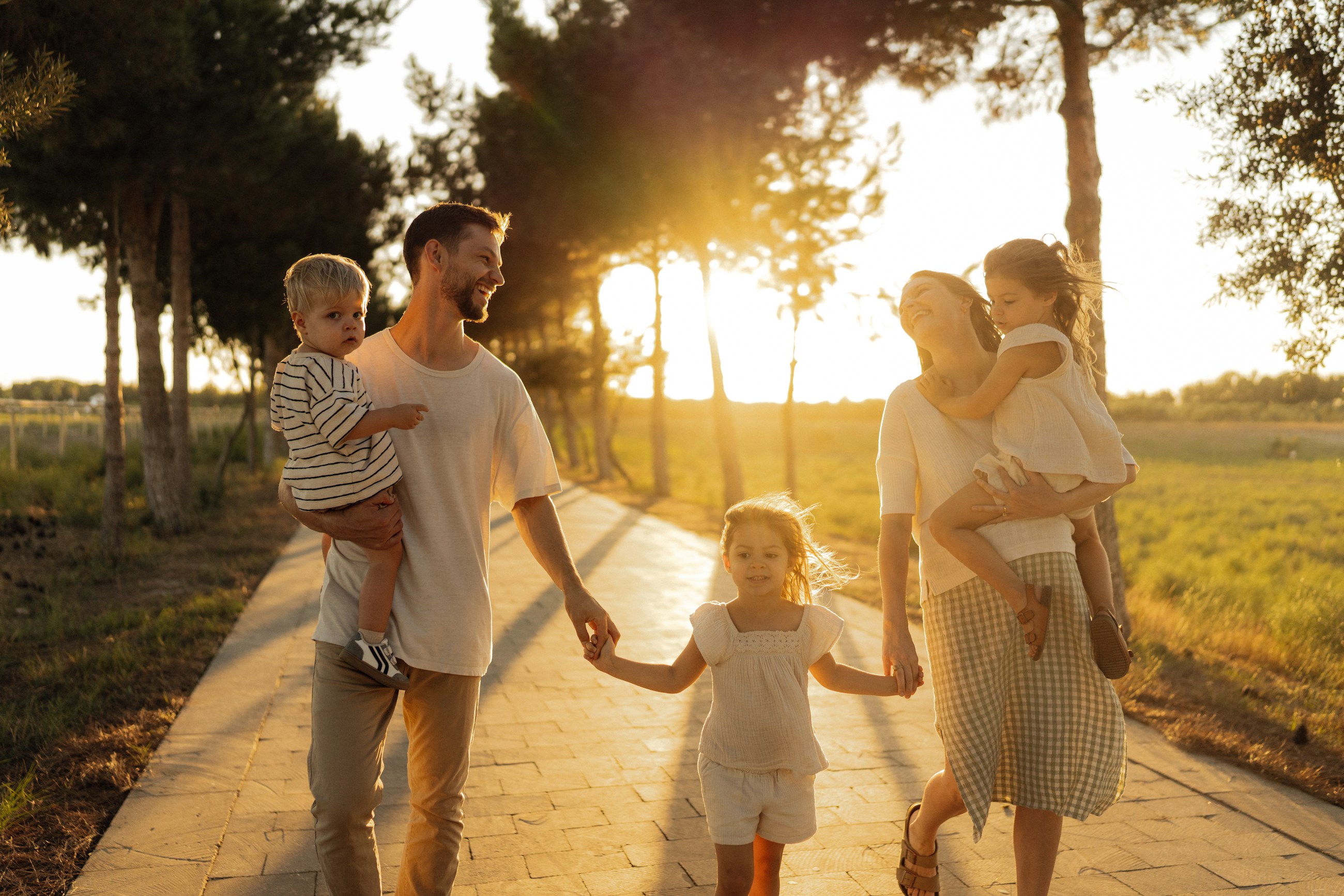 A father walks ahead carrying his two youngest children while his wife follows holding their daughter's hand, golden hour on a tree-lined path